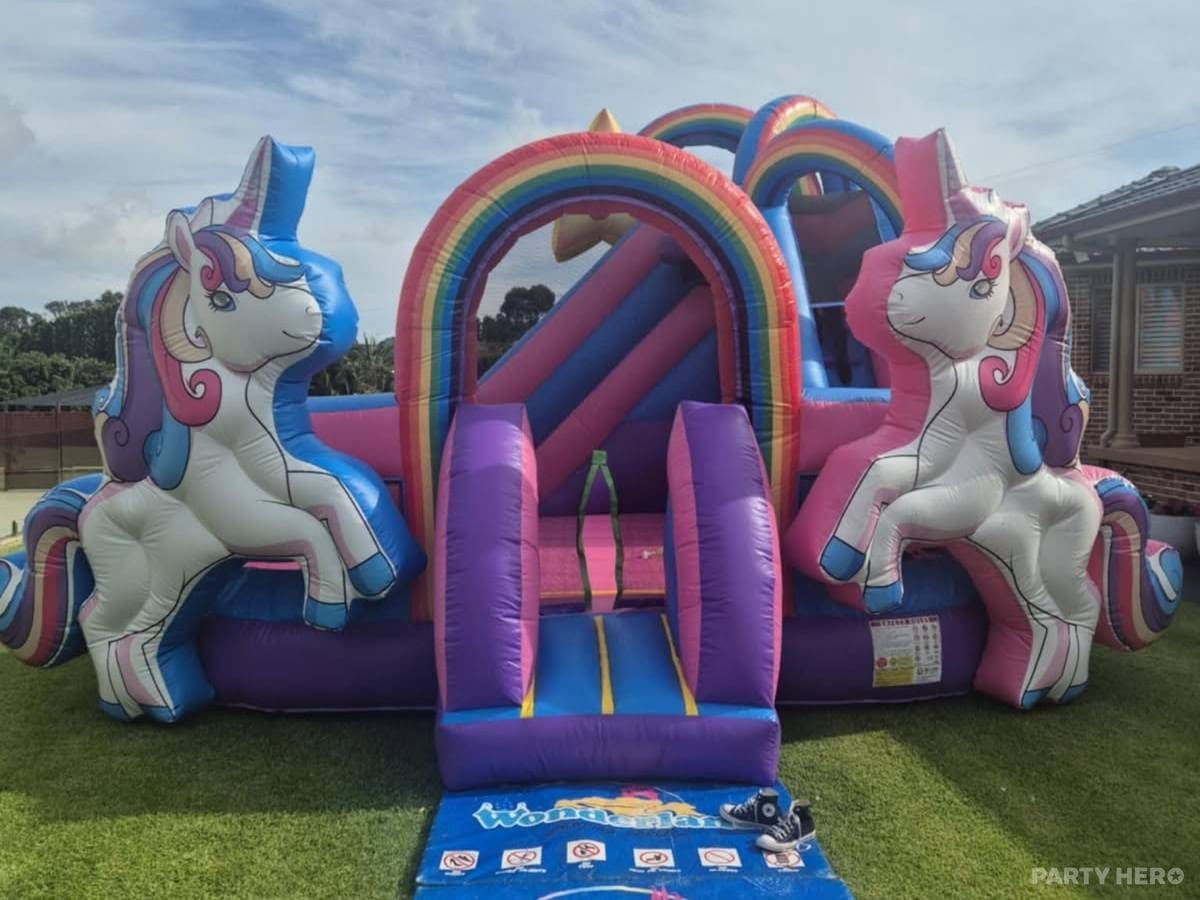 Pink and purple unicorn castle with a double slide and rainbow arch, set up on a lawn