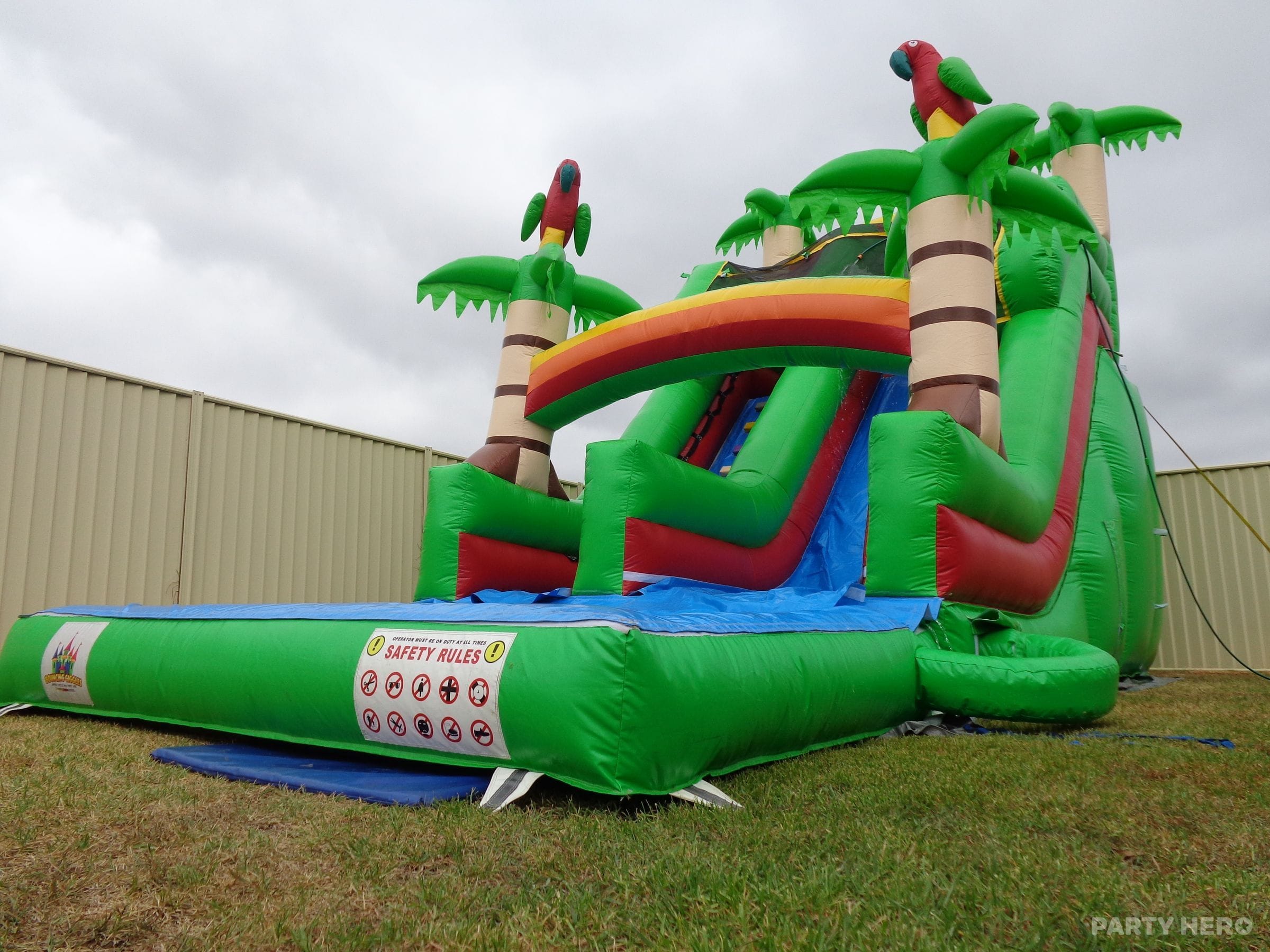 Green Jungle water slide with palm trees and parrots, set up in a fenced backyard