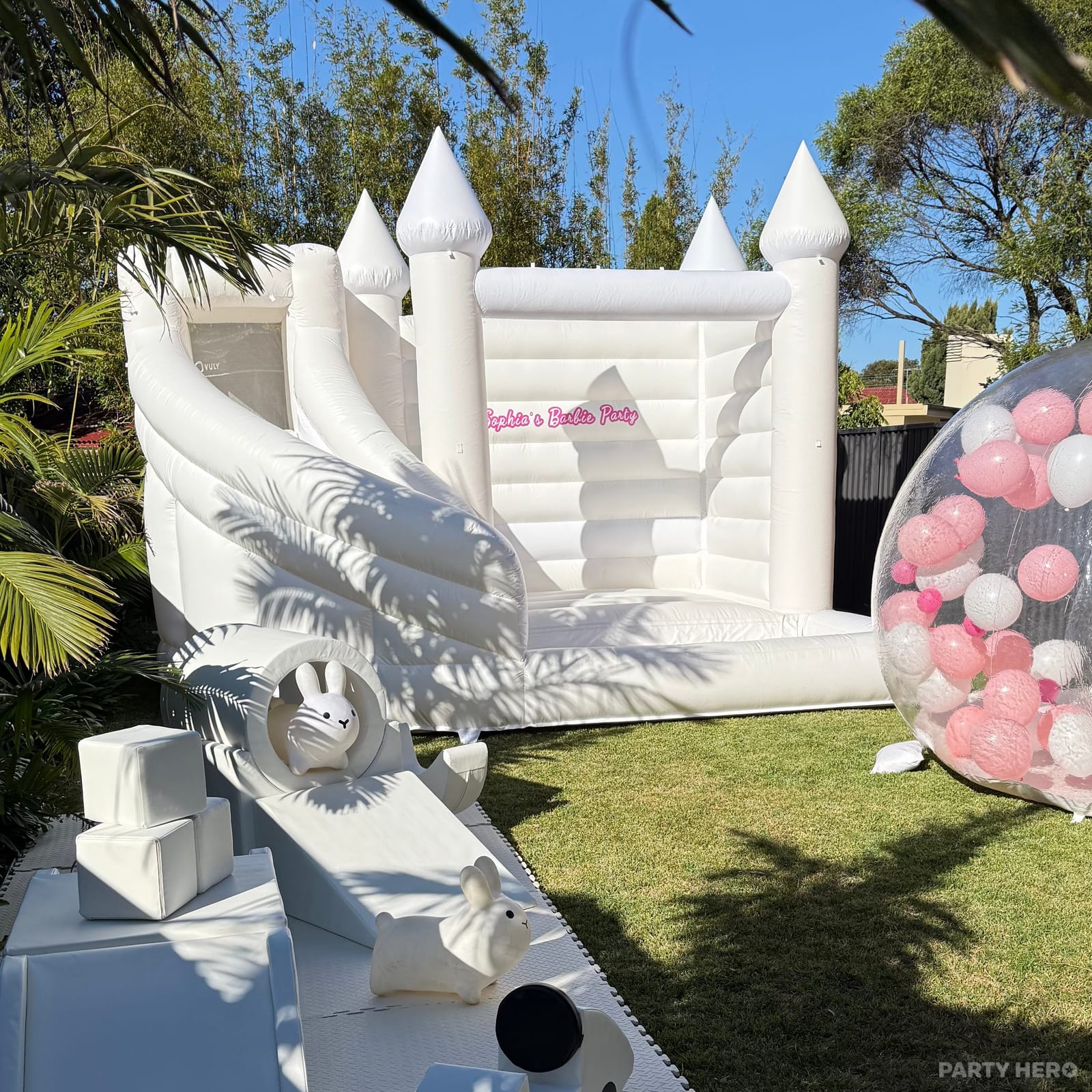 White bounce house with a curved slide and a balloon-filled bubble dome on a lawn